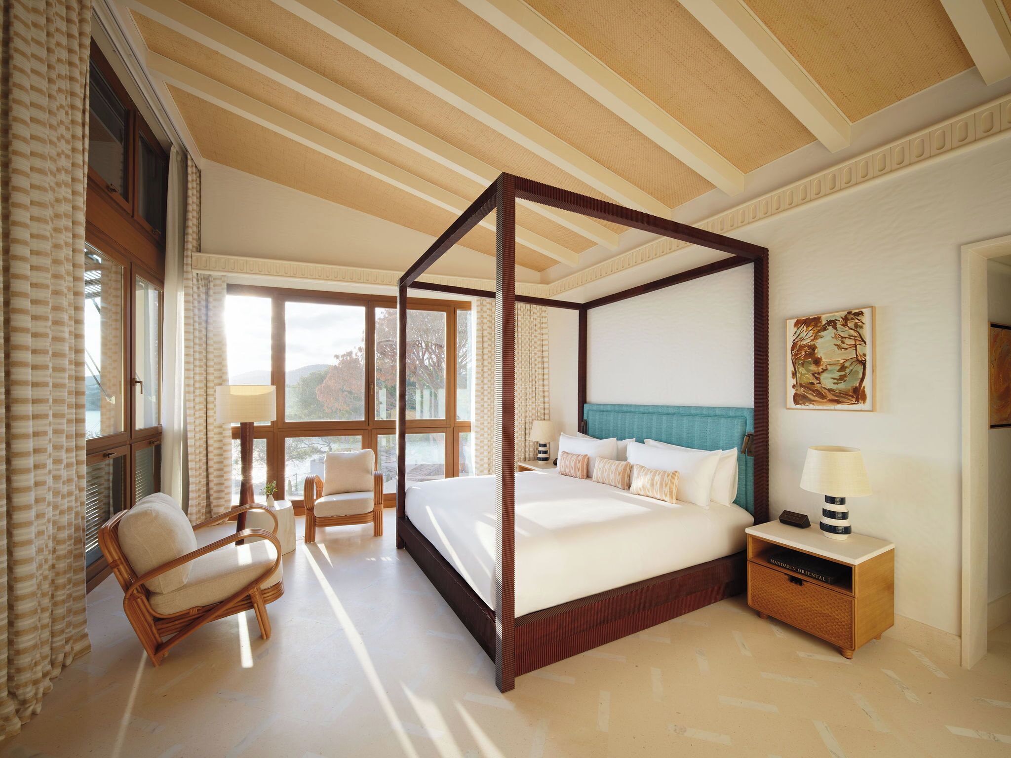 A four-poster bed with a view of the outdoors, a wicker chair, a bedside table with a lamp, and a painting on the wall.