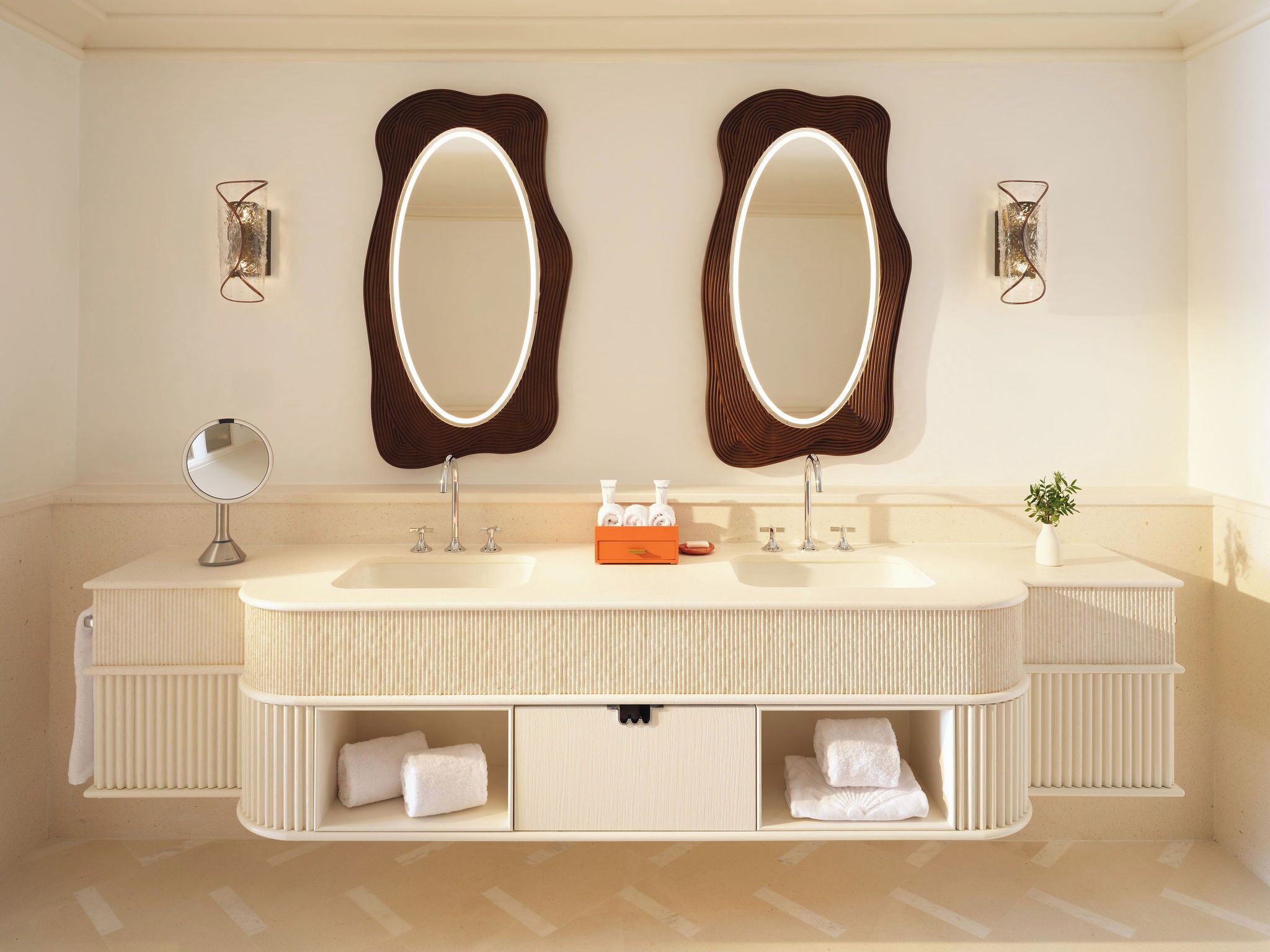 A bathroom with two large mirrors, a sink with two faucets, a small round mirror, and a potted plant.