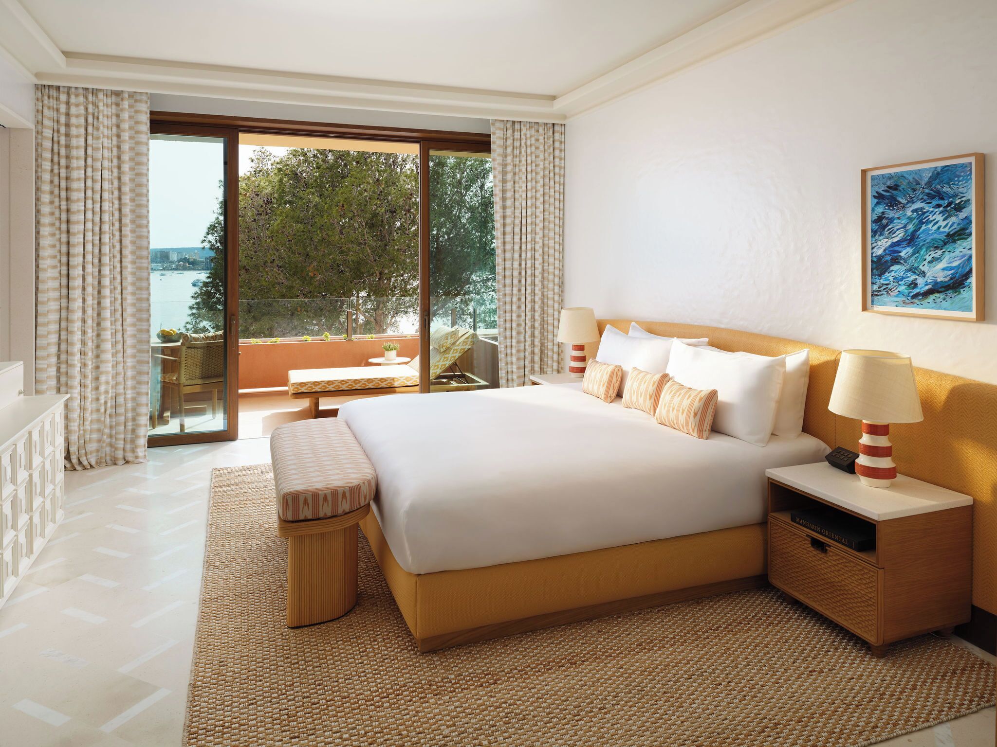 A hotel room with a large bed, a bedside table, a lamp, a view of the outdoors, and a painting on the wall.