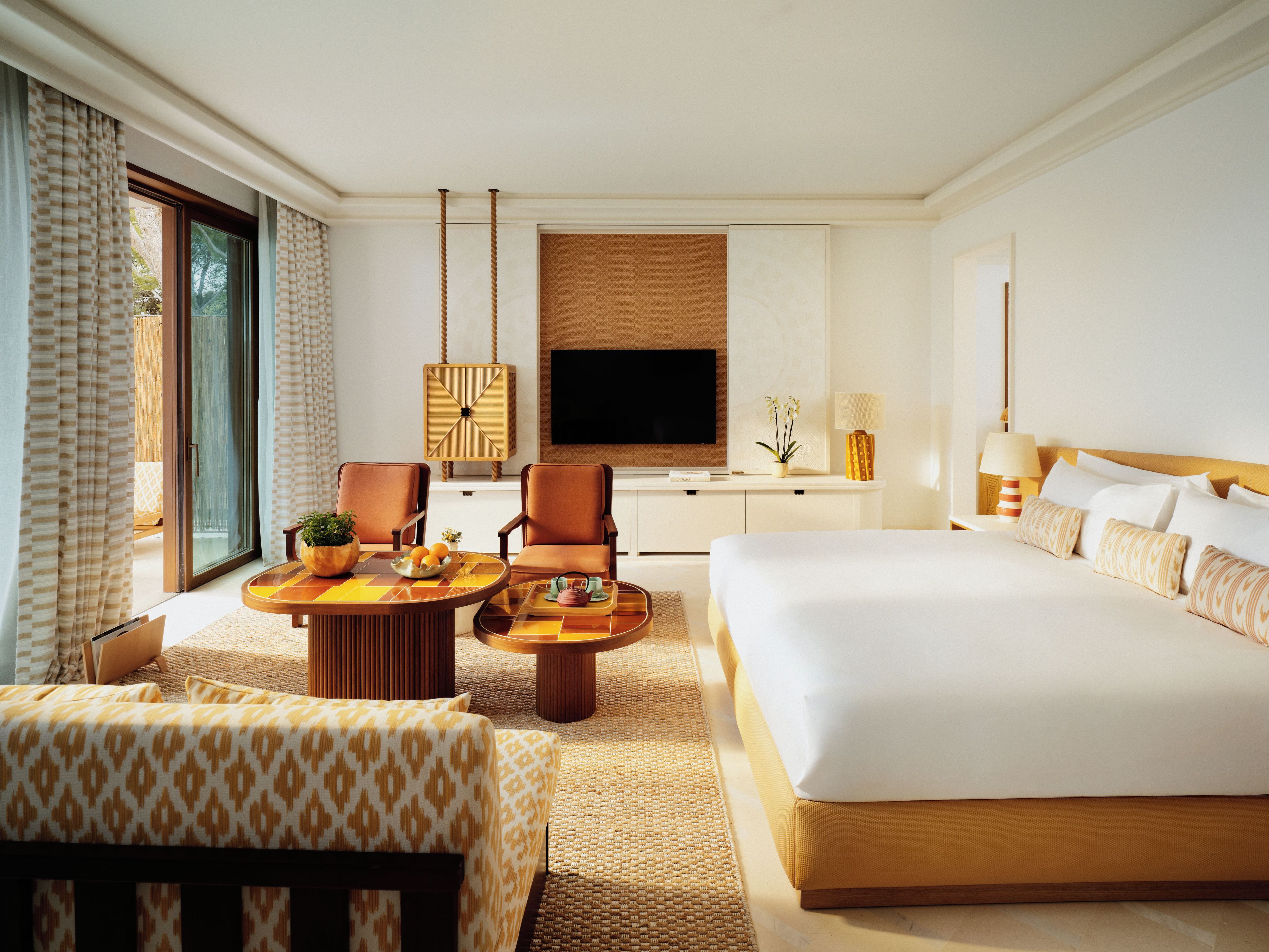 A hotel room with a large bed, two armchairs, a small table with a tray of fruit, a television, and a window with curtains.