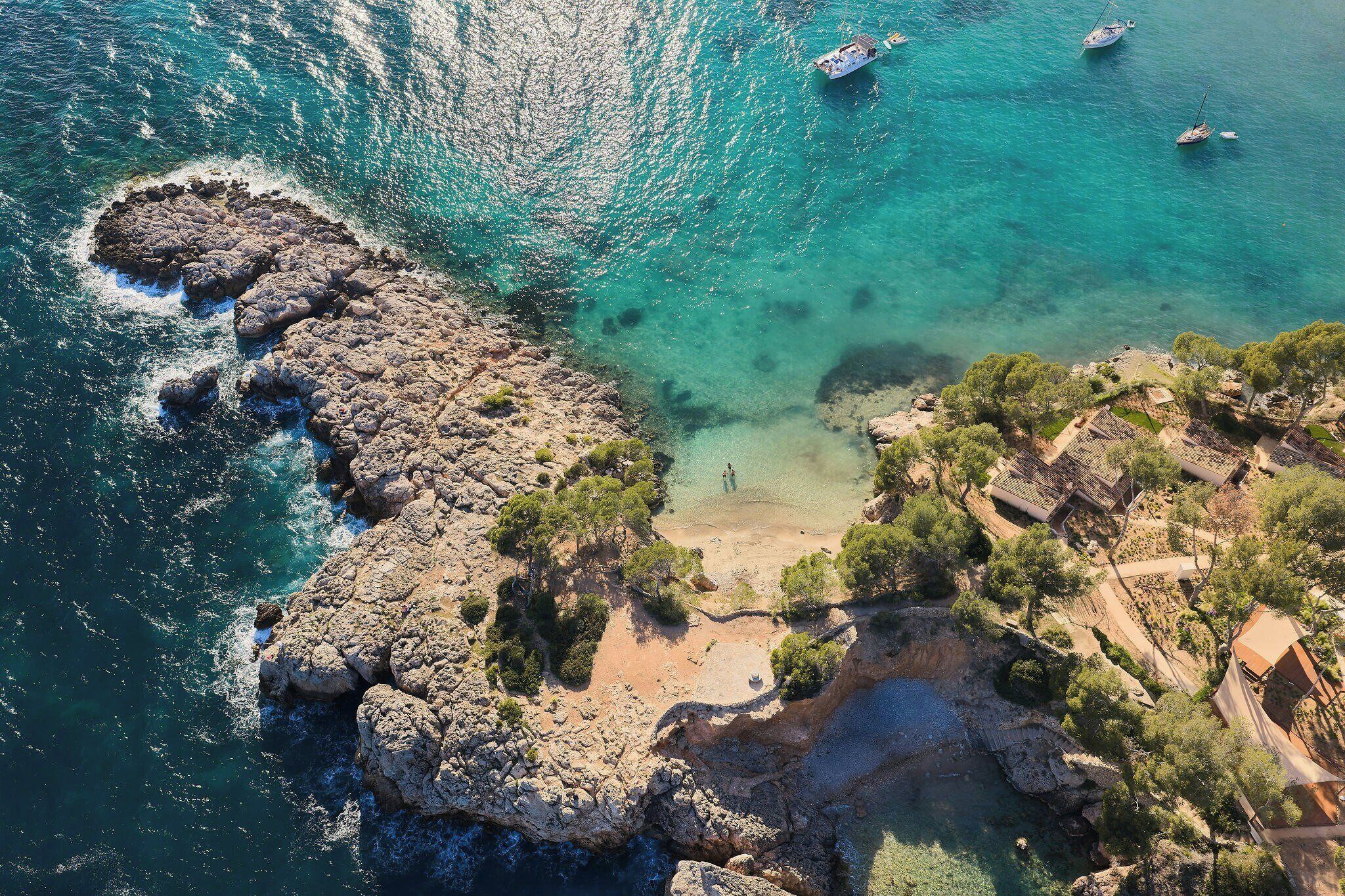 An aerial view of a coastal area with rocky terrain, clear water, boats, and sparse vegetation.