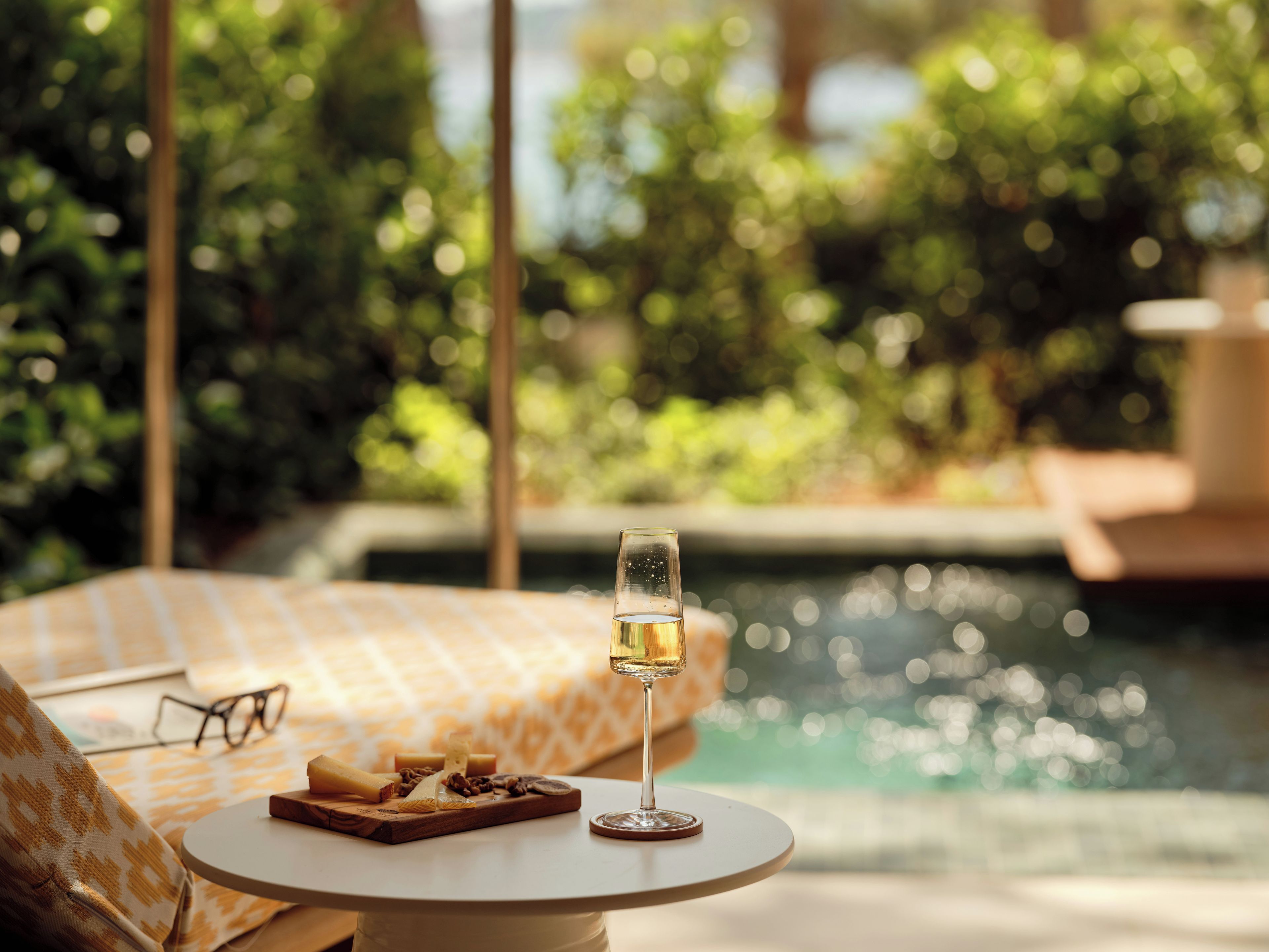 Un verre de vin blanc sur une table ronde, accompagné d'une planche de fromages, de verres et d'un livre, près d'une piscine avec un arrière-plan verdoyant flou.