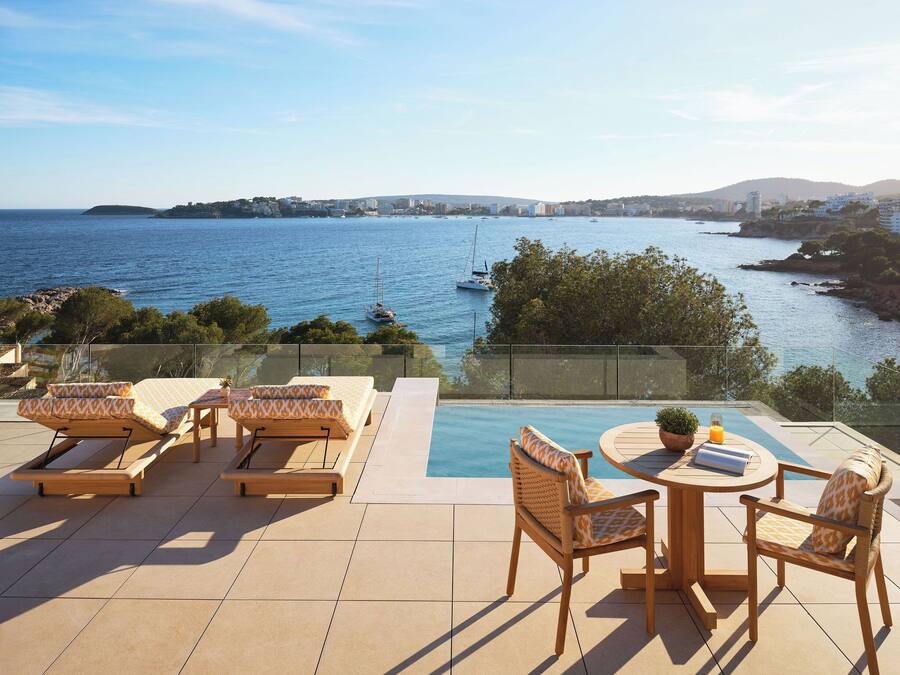 Una terraza con piscina, sillones de descanso y una mesa con sillas, con vistas a la costa, barcos y edificios.