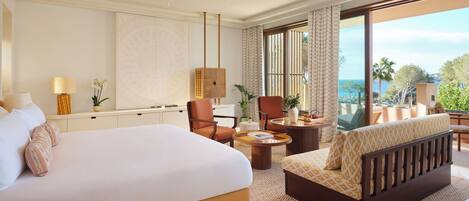 A hotel room with a large bed, a sofa, a chair, a small table, and a view of the sea.