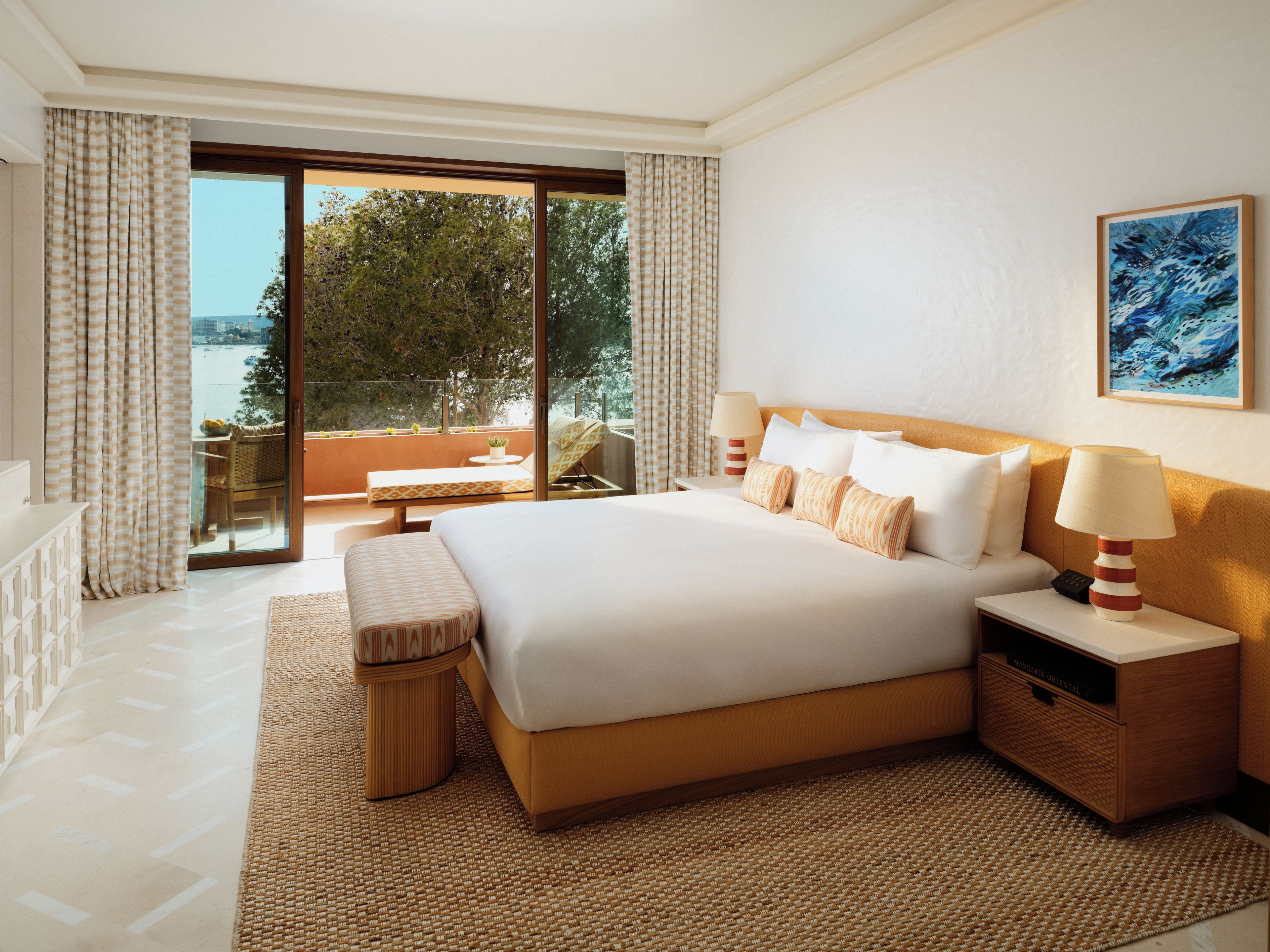 A hotel room with a large bed, a bedside table with a lamp, a small table with a chair, and a view of the sea through the window.