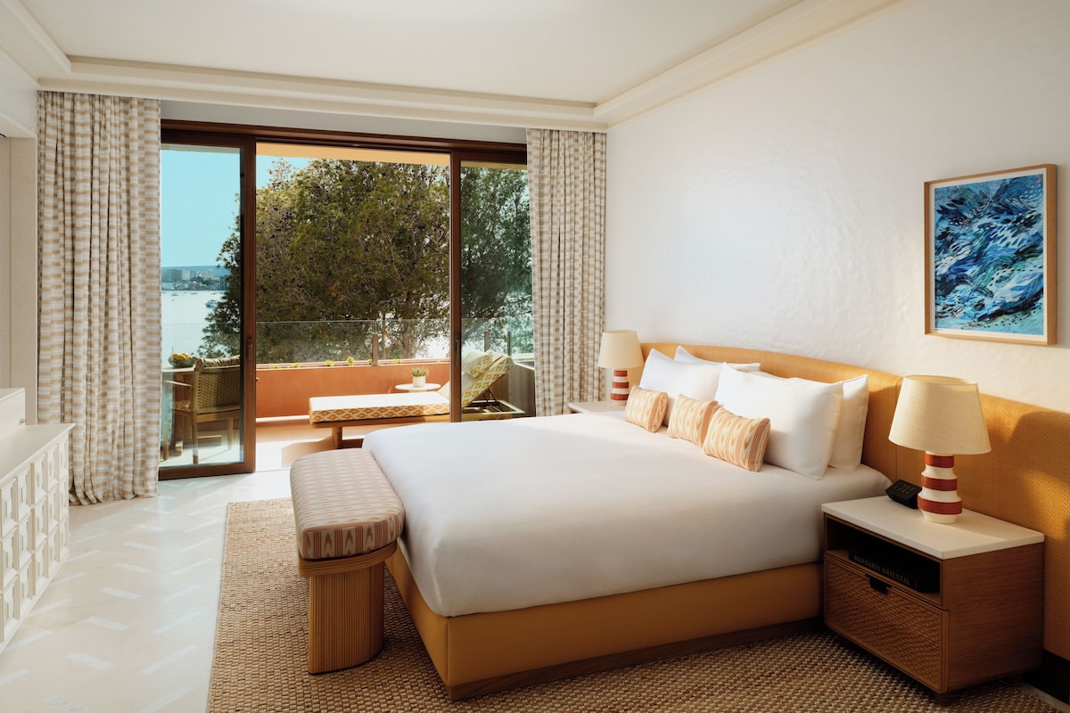 A hotel room with a large bed, a bedside table with a lamp, a small table with a chair, and a view of the sea through the window.