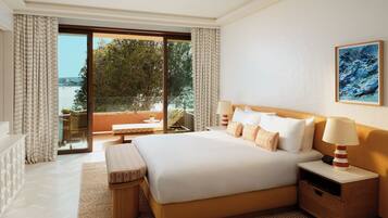 A hotel room with a large bed, a bedside table with a lamp, a small table with a chair, and a view of the sea through the window.