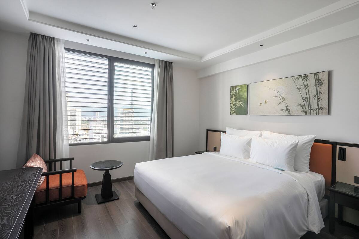 A hotel room with a large bed, a small table, a chair, and a window with blinds.