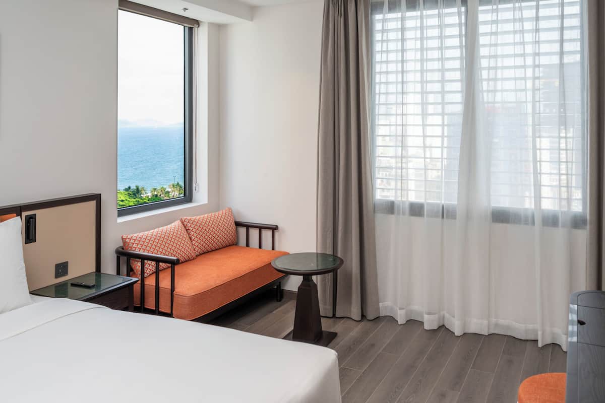 A hotel room with a bed, a sofa with orange cushions, a small table, and a view of the ocean through a window.