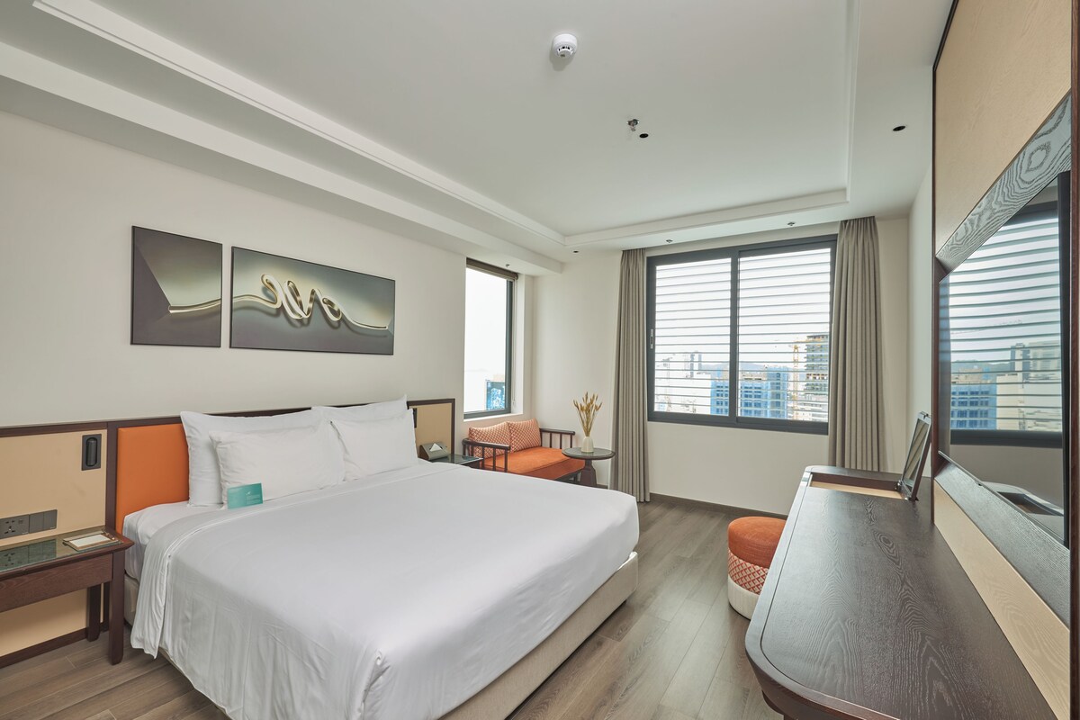 A hotel room with a large bed, a desk, a chair, a window with a view of buildings, and artwork on the wall.