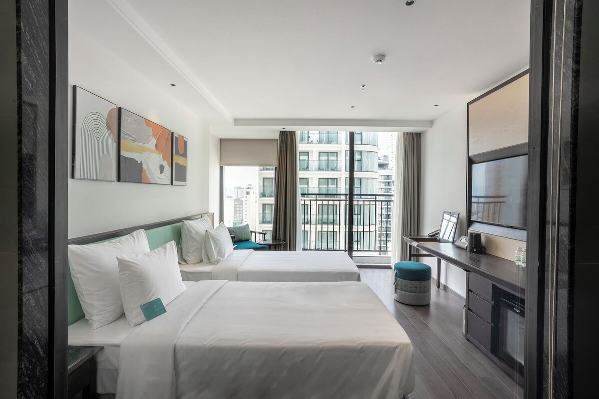 A hotel room with two beds, a desk, a TV, and a large window with a city view.