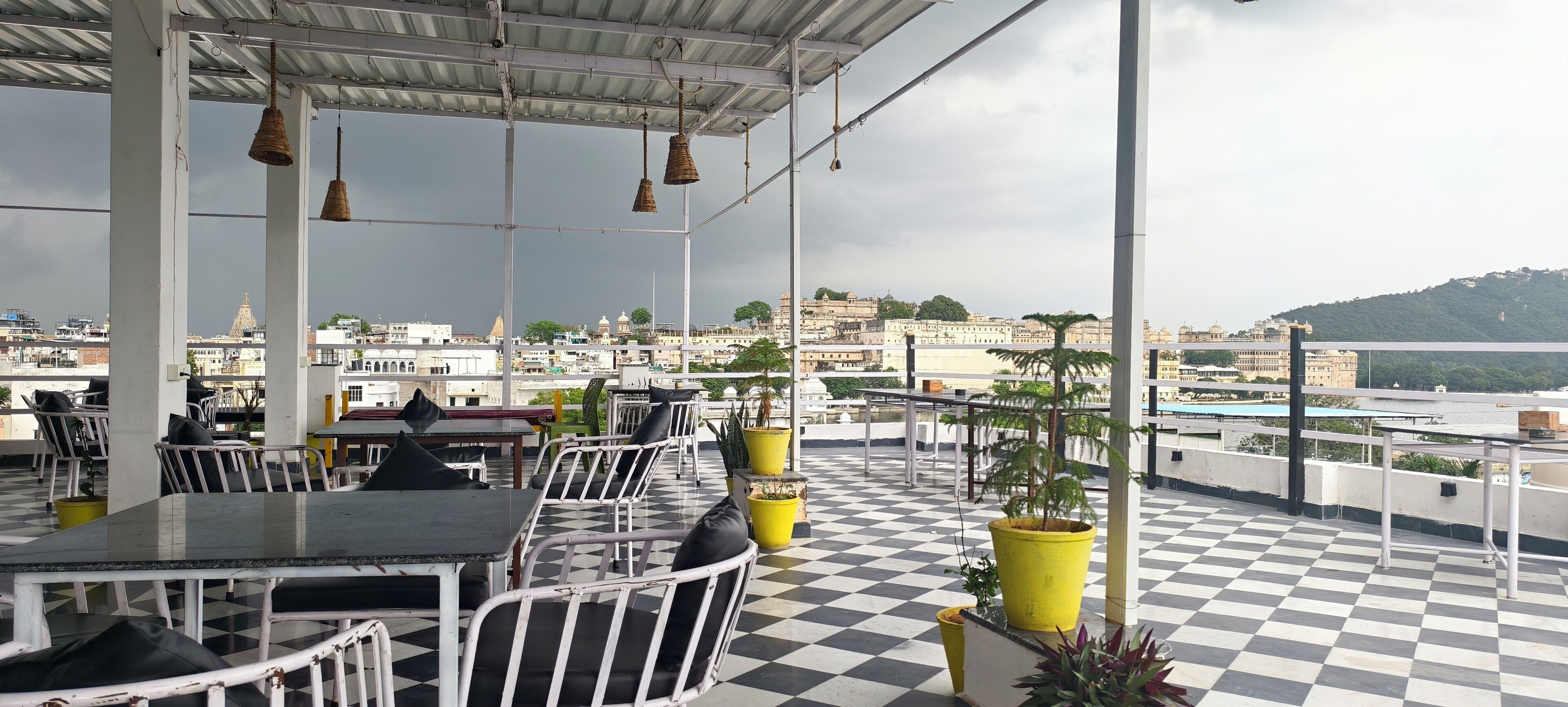 An outdoor terrace with checkered flooring, metal tables and chairs, hanging wicker lamps, and potted plants.