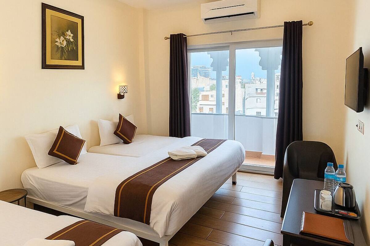 Ein Hotelzimmer mit zwei Betten, einem Schreibtisch, einem Sessel, einem Deckenventilator, einer Klimaanlage, einem Fernseher und einem Fenster mit Vorhängen.