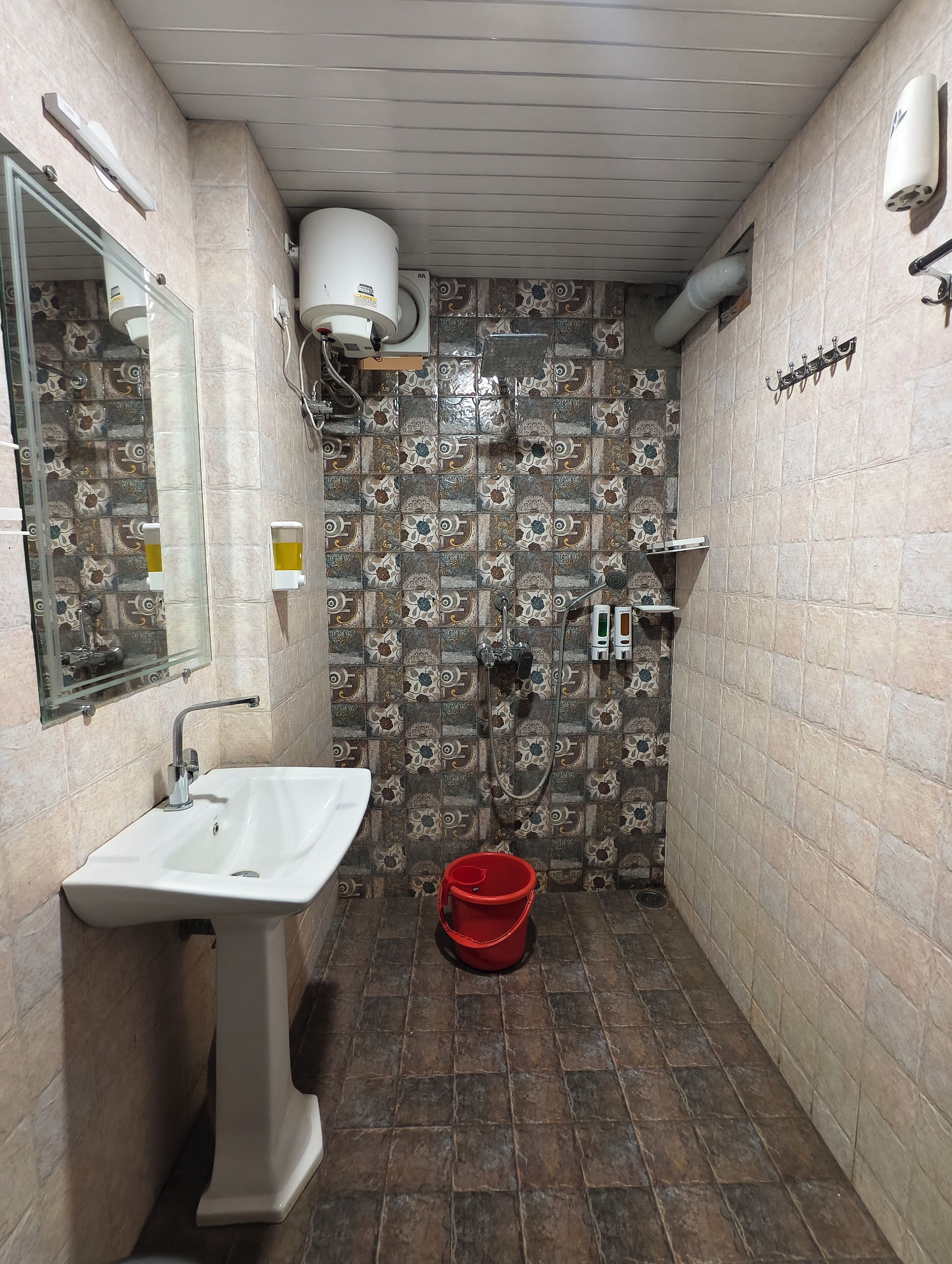 A bathroom with a pedestal sink, a shower with a handheld showerhead, a red bucket, and patterned tiles on the walls.