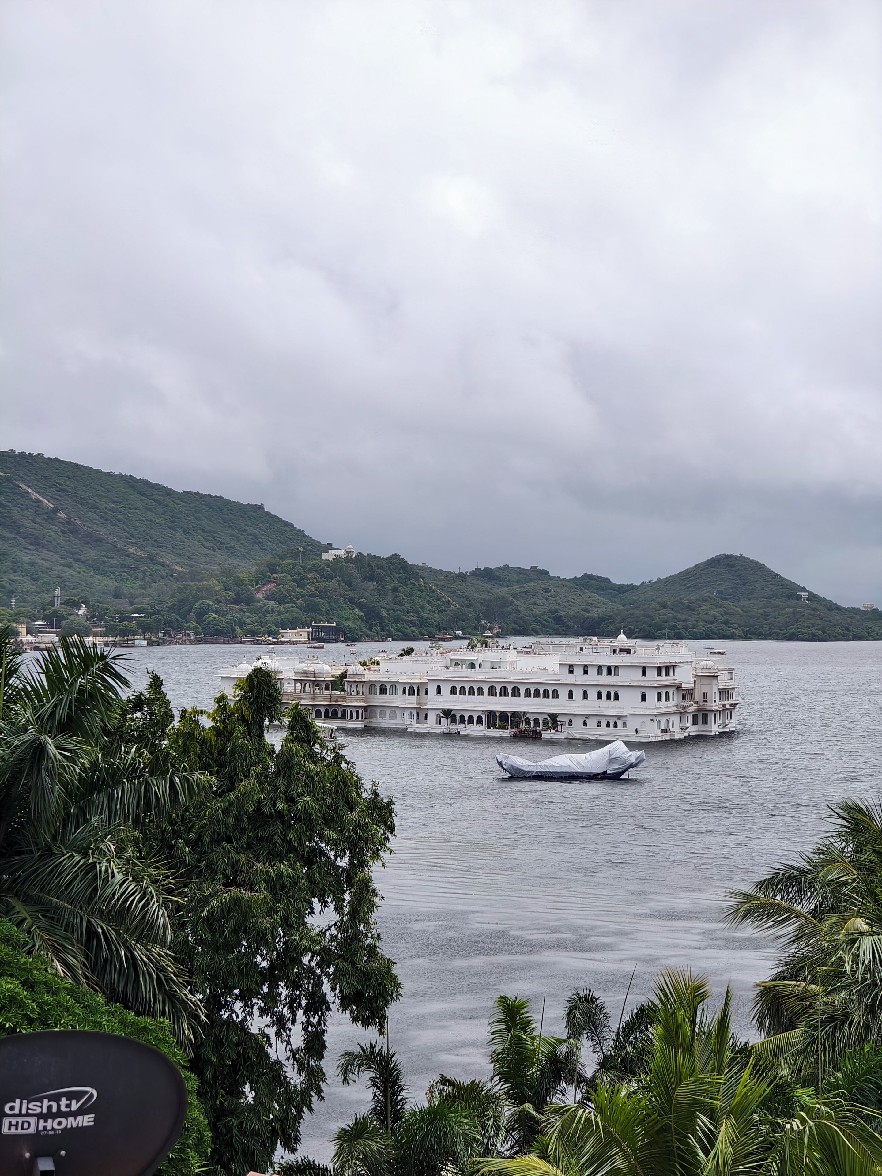 A large, multi-story building with a white facade, situated on a body of water, surrounded by greenery and hills.