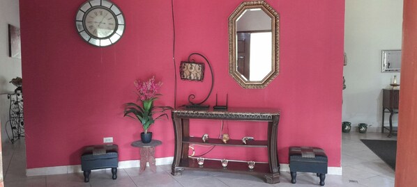 Una habitación con pared roja, un reloj redondo, un espejo, una consola decorada con objetos y dos taburetes pequeños.