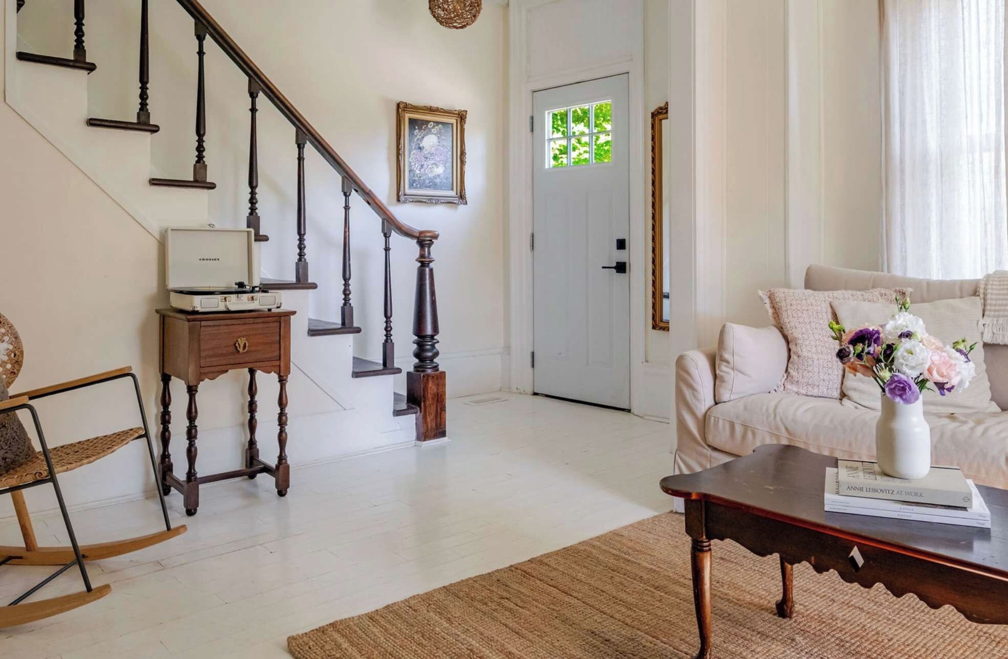 A living room with a staircase, a white door, a sofa, a rocking chair, and a coffee table with a book.