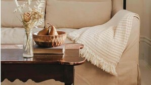 A living room with a beige sofa, a table with a vase of dried flowers, a bowl of pears, and a lamp.