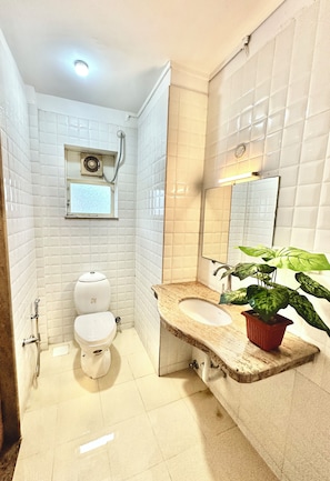 Une salle de bain avec des murs recouverts de carreaux blancs, des toilettes, un lavabo avec un miroir et une plante en pot.
