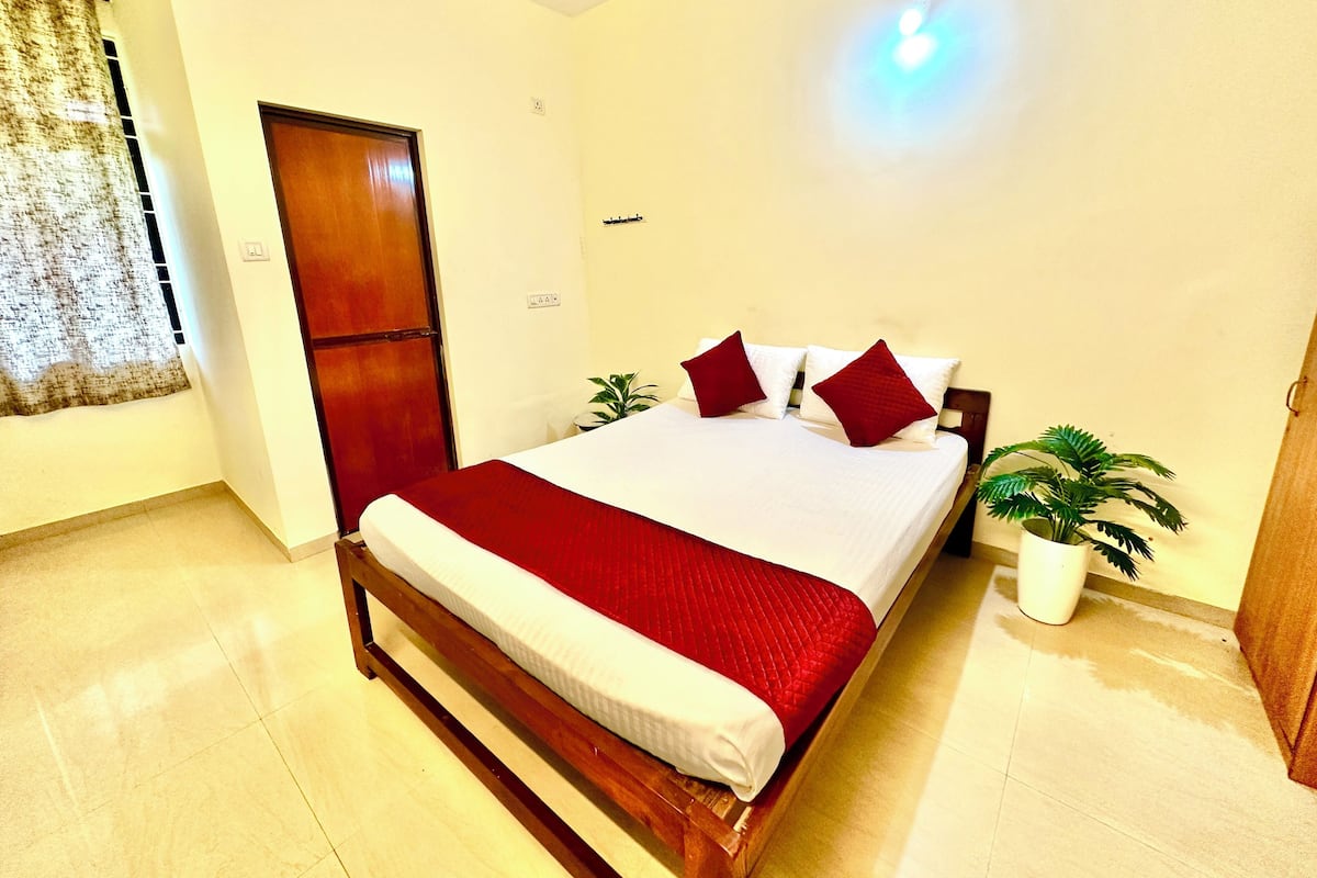 A neatly made bed with a wooden frame and red bedding, a potted plant, and a door to the left.