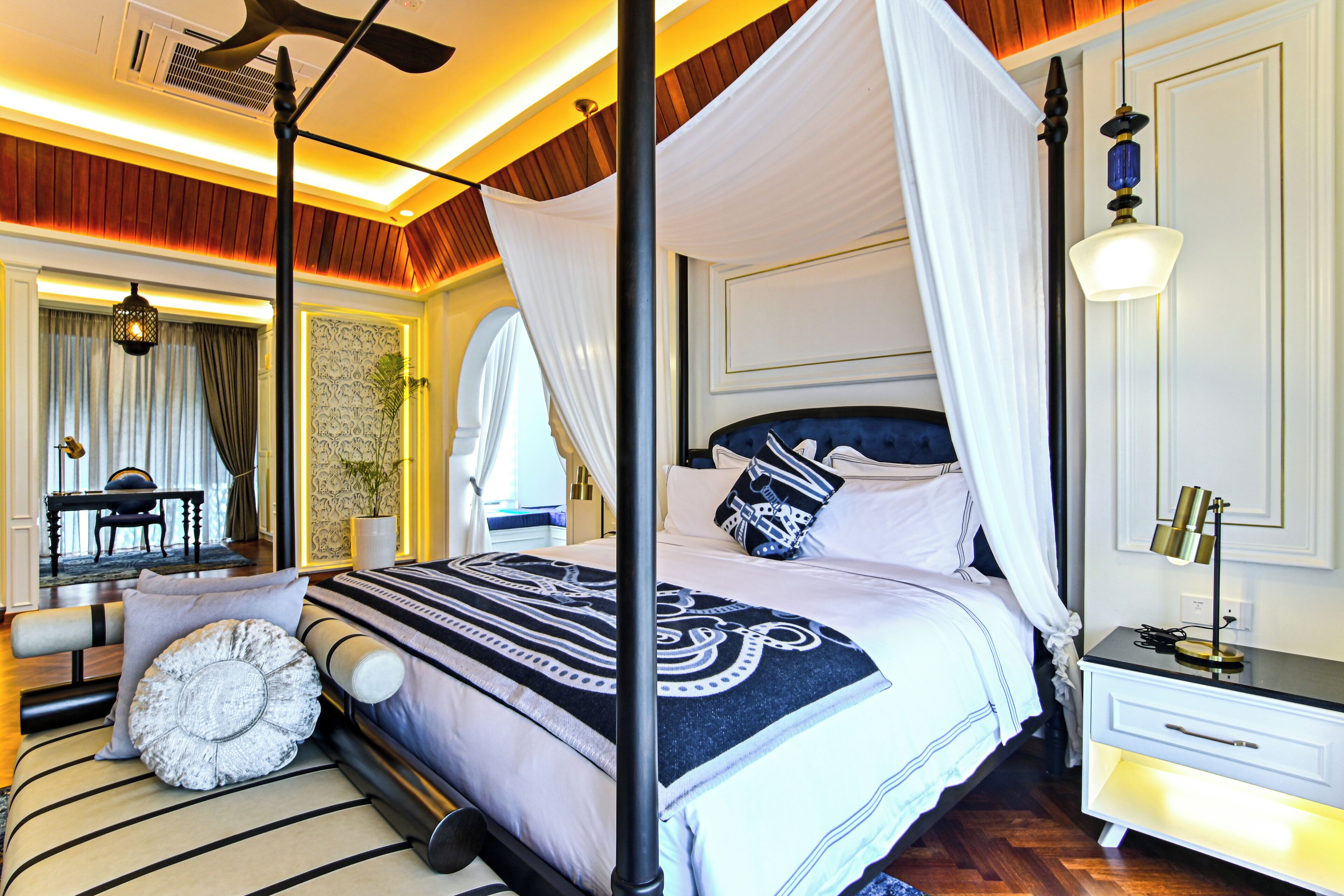 A four-poster canopy bed with a white and blue patterned bedspread, a bedside table with a lamp, and a view of a dining area with a table and chairs.