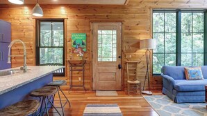 A wooden cabin interior with a kitchen, bar stools, a blue sofa, and large windows. - Cottage at Deer Ridge Farm (Charlottesville)