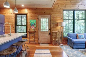 A wooden cabin interior with a kitchen, bar stools, a blue sofa, and large windows.