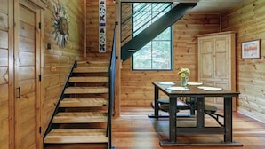 A wooden cabin interior with a staircase, a dining table with chairs, and a window with a view of trees. - Cottage at Deer Ridge Farm (Charlottesville)