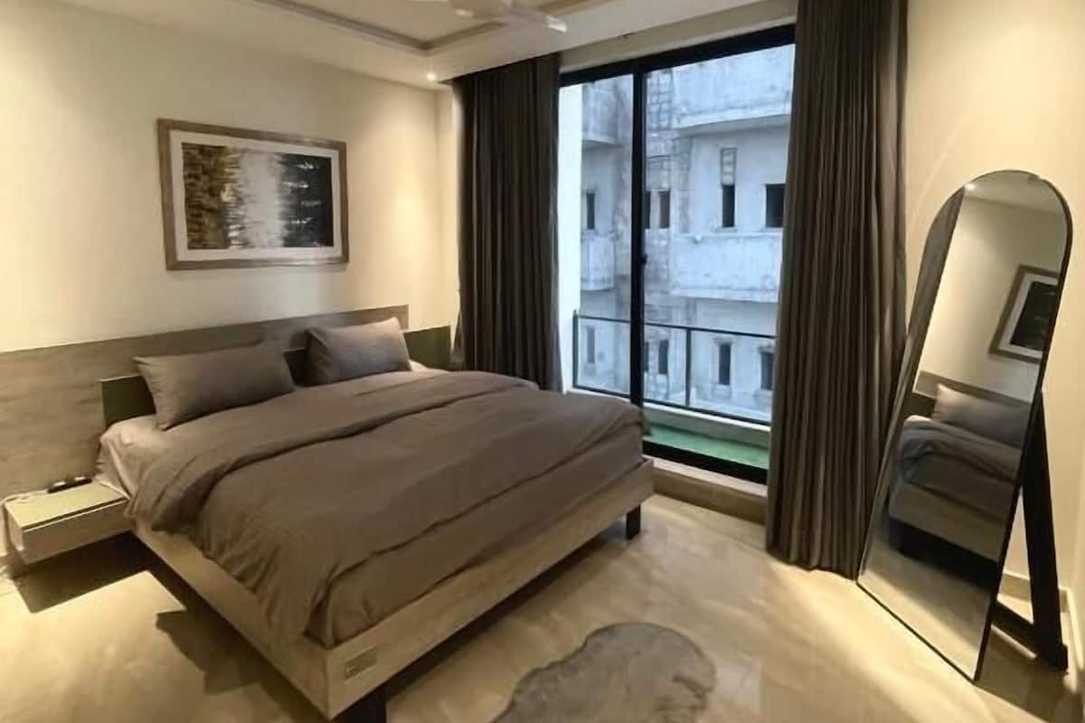 Un dormitorio moderno con una cama grande, un ventilador de techo, una mesita de noche, un cuadro enmarcado, un ventanal con cortinas y un espejo.