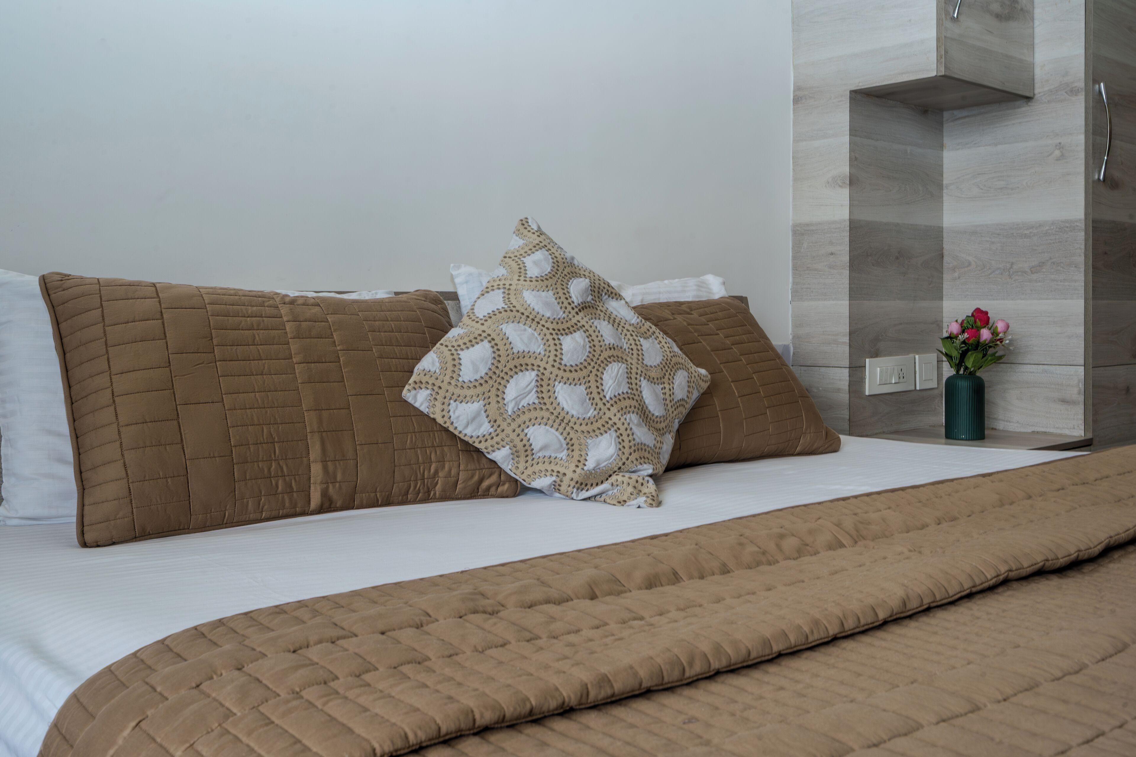 A neatly made bed with brown bedding and a patterned pillow, a vase with flowers, and a modern bathroom with a wooden vanity and a towel rack.