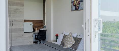 A modern bedroom with a bed, a desk, and a chair. There is a painting on the wall and a window with a view of greenery.