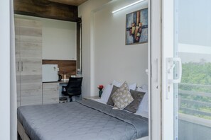 A modern bedroom with a bed, a desk, and a chair. There is a painting on the wall and a window with a view of greenery. - Stay Studio Hotel & Residences (Gurugram)