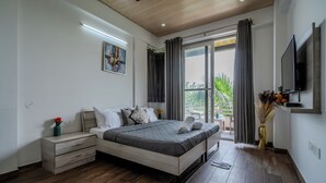 Ένα σύγχρονο υπνοδωμάτιο με ξύλινο δάπεδο, ένα κρεβάτι με γκρι κλινοσκεπάσματα, ένα κομοδίνο, ένα βάζο με αποξηραμένα φυτά και μια τηλεόραση στον τοίχο.