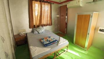 A small, single-bed room with a green floor, a wooden bed frame, a nightstand, a bedside table, a window with curtains, and a door.
