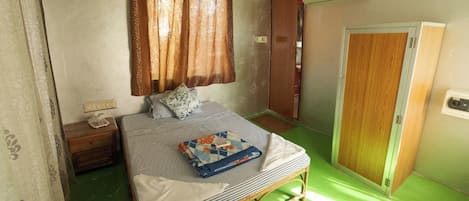 A small, single-bed room with a green floor, a wooden bed frame, a nightstand, a bedside table, a window with curtains, and a door.