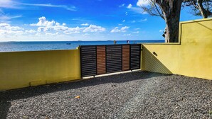 A yellow wall with a black gate leading to a pebbled area by the sea. - Charming 2-bedroom villa with WiFi, AC in pleasant Vieux Grand Port (Vieux Grand Port)