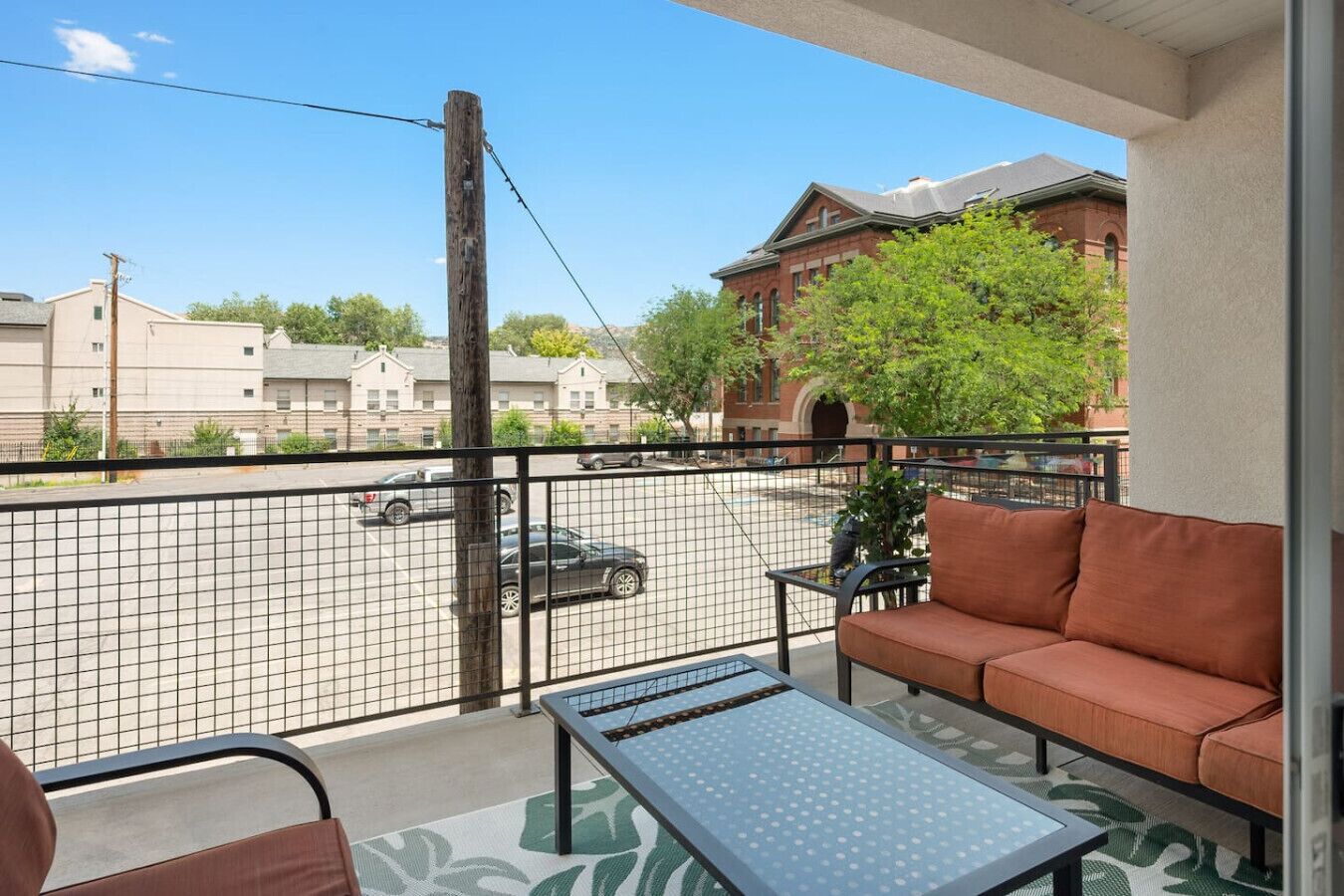 A balcony with a sofa and a table, a view of a parking lot, and a building in the background.
