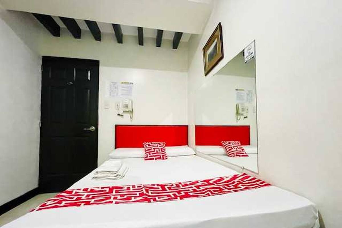A hotel room with a bed, a black door, a red headboard, a mirror, and a white wall.