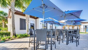 An outdoor patio area with wicker tables and chairs, large blue umbrellas, and a building with a tiled roof. - Pool & Lazy River: Windsor House 9 Mi to Disney! (Kissimmee)