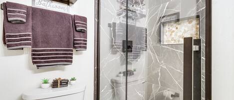 Un baño moderno con ducha de vidrio, inodoro y lavamanos. Hay dos toallas colgadas en la pared y un estante con artículos de aseo.