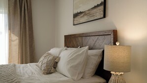 Een net opgemaakt bed met witte lakens en een plaids, een houten hoofdbord, een bedlamp en een lijst met een landschapsafbeelding aan de muur.