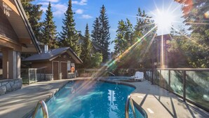 Une piscine où le soleil filtre à travers les arbres, entourée de bâtiments et sous un ciel dégagé.