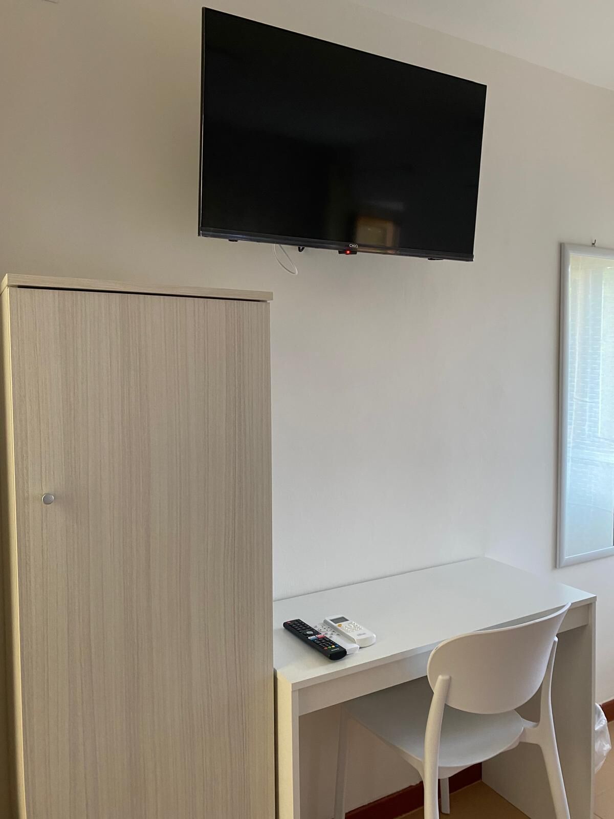 Quarto com uma escrivaninha branca, uma cadeira branca, uma televisão fixada na parede e um guarda-roupa.