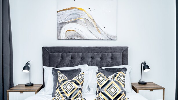 A bedroom with a grey upholstered headboard, a white bedspread, and two bedside tables with lamps. There is a wall-mounted abstract painting above the bed. - Hilltown by Central Serviced Apartments (Dundee)