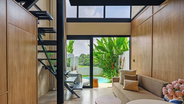 A modern interior with a staircase, a sofa, a small round table, and a view of a swimming pool and greenery outside. - Exhale Villa by Betterplace (Canggu)