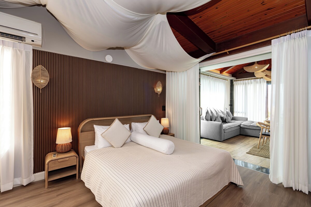 Een hotelkamer met een groot bed, een houten plafond, een wandairconditioner en uitzicht op een aangrenzend woongedeelte.