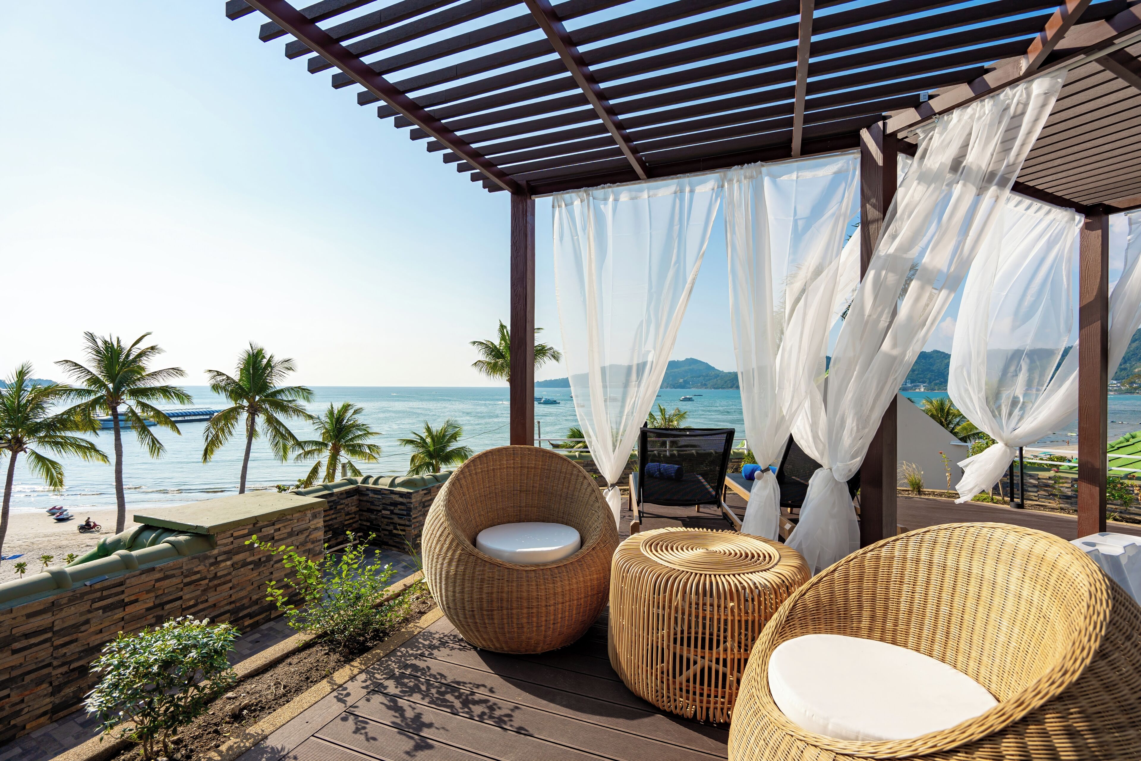A beachside patio with wicker furniture, a view of the ocean, and a wooden pergola.
