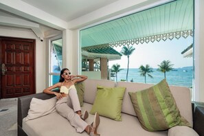 Een kamer met een groot raam dat uitkijkt over een strand, een houten deur en een bank met kussens.