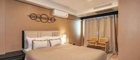 A hotel room with a large bed, a bedside table, a lamp, a chair, and an air conditioning unit.