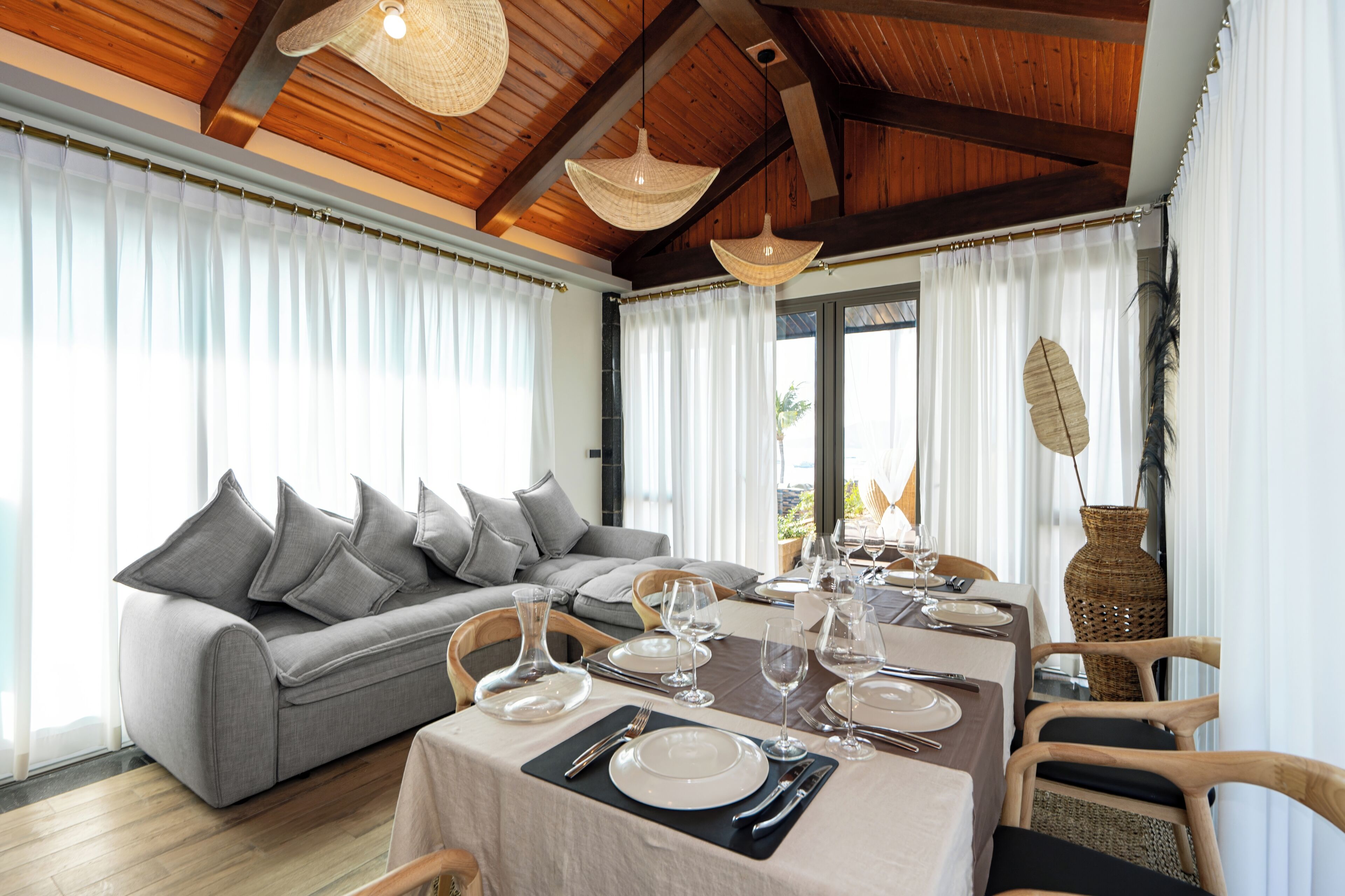 A dining area with a table set for four, a grey sofa, and a large window with curtains.
